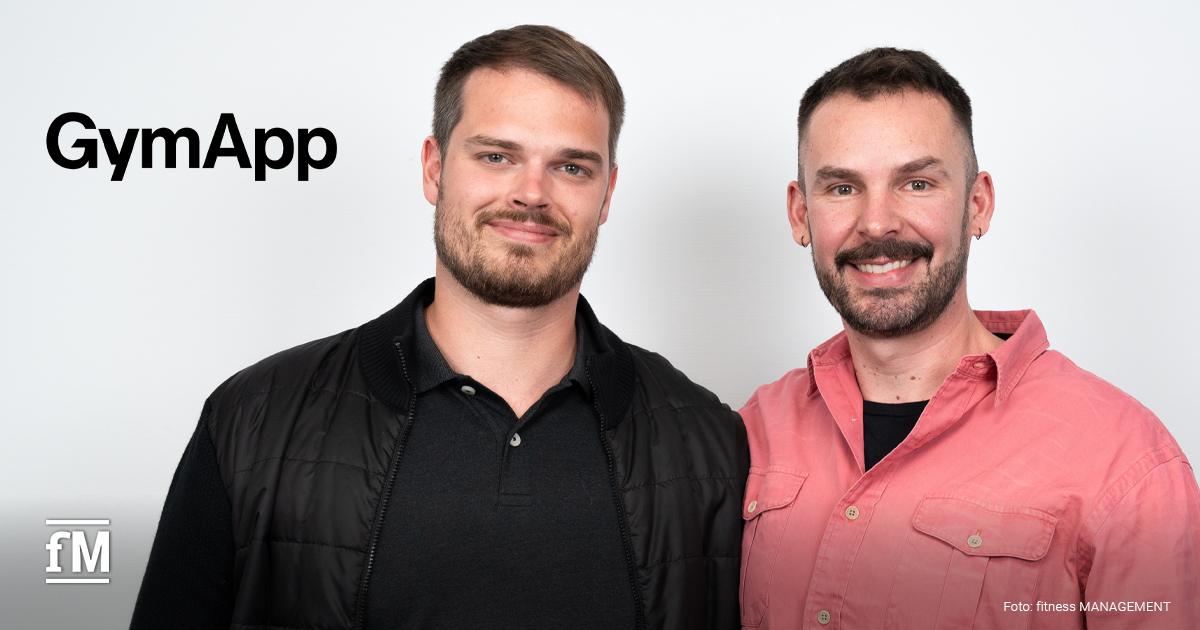 Zwei Männer stehen nebeneinander vor einer weißen Wand. Links trägt der Mann (Robin Lausch) eine schwarze Jacke und ein dunkles Poloshirt, rechts trägt der Mann (Janosch Marx) ein rosafarbenes Hemd. Links oben ist das schwarze GymApp-Logo zu sehen.