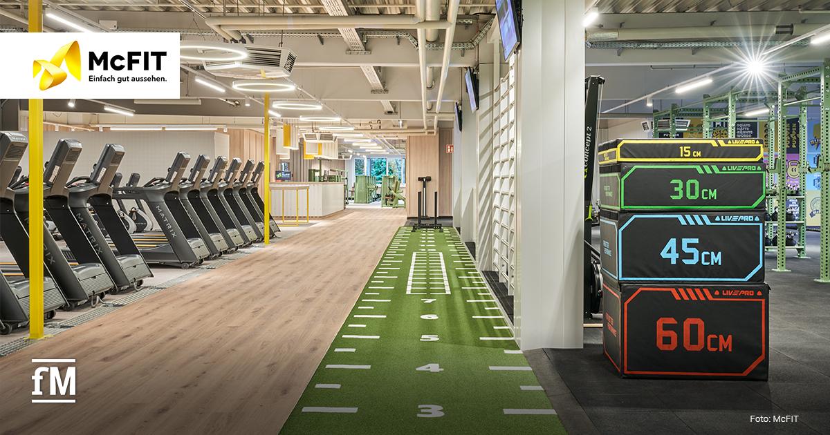 Grüne Kunstrasenbahn mit aufgestapelten Sprungboxen und Laufbändern in einem Fitnessstudio. Links befindet sich das McFIT-Logo.