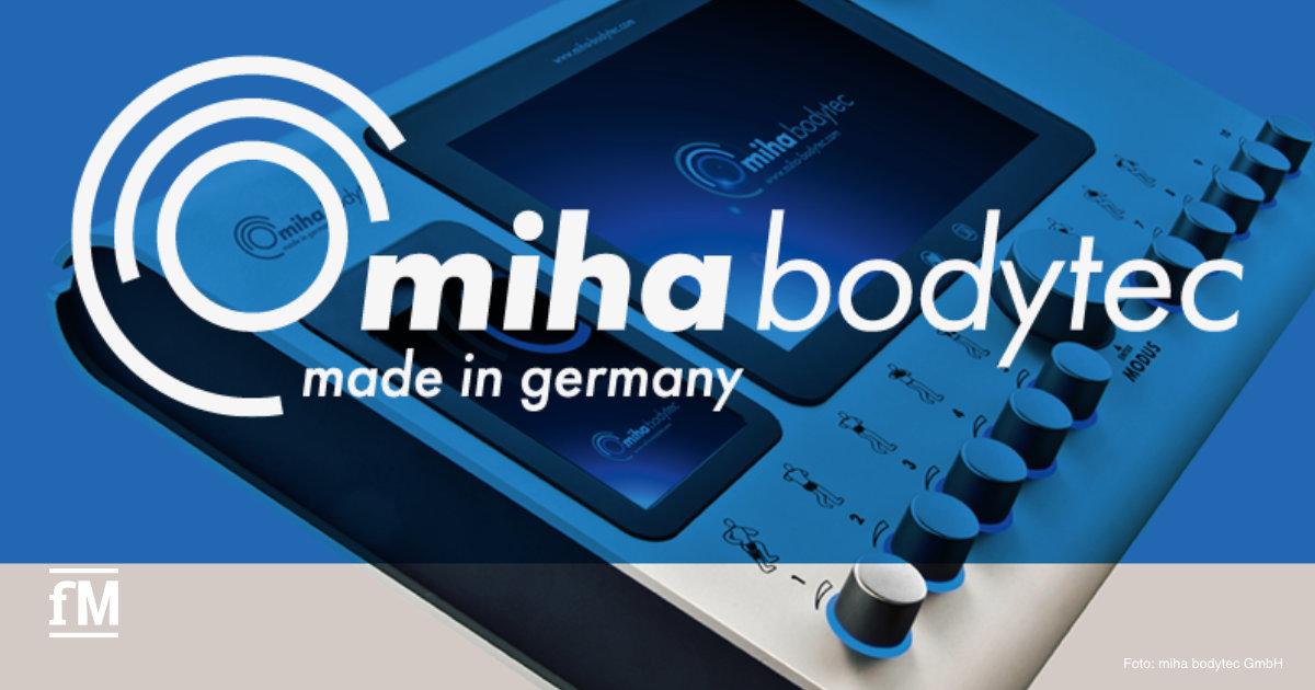 Nahaufnahme eines EMS-Bediengeräts mit Display und mehreren Drehknöpfen; darüber groß eingeblendetes Logo 'miha bodytec – made in germany'; links unten kleines fM-Logo; blau-graue Farbgebung.