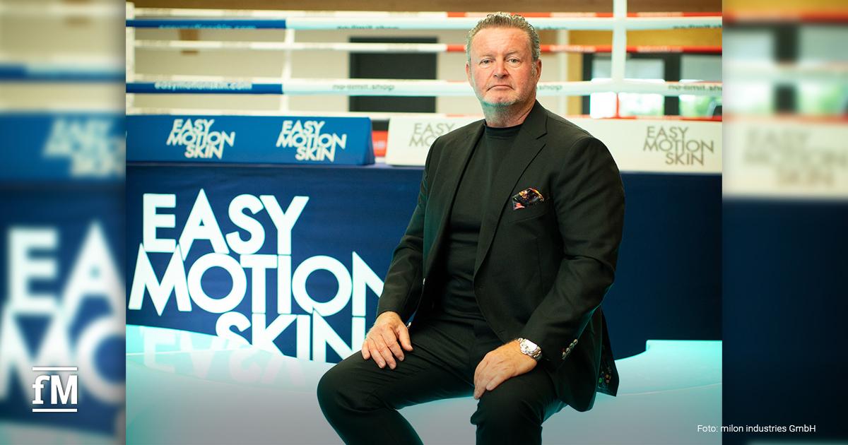 Mann (Christian Jäger) in schwarzem Anzug sitzt vor Boxring mit der Aufschrift EasyMotionSkin.