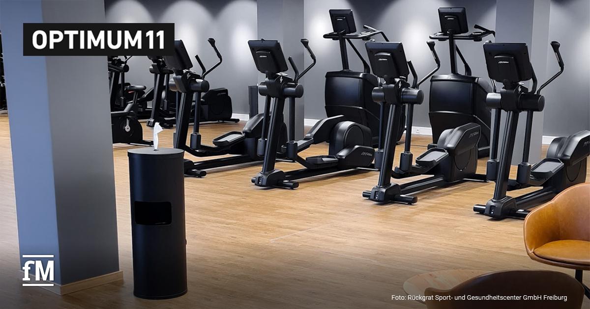 In einem modernen Fitnessstudio stehen Cardiogeräte und ein Spender für Desinfektionstücher. Oben links ist das Logo von Optimum11.