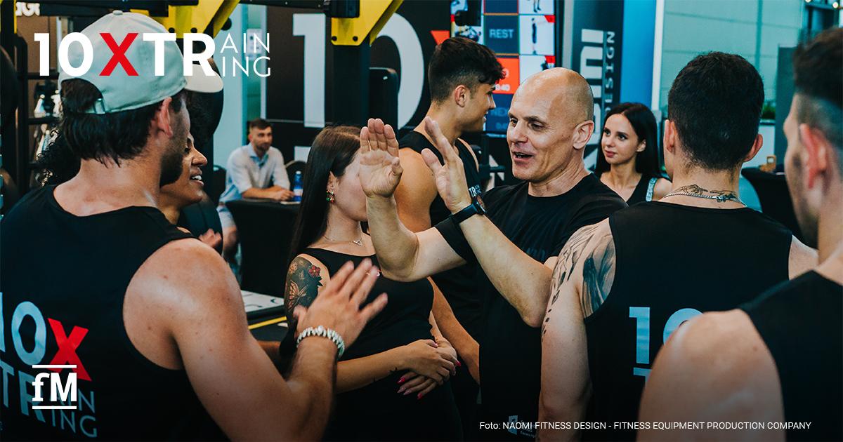 Gruppe von Menschen in Sportkleidung gibt sich im Fitnessstudio High-Fives. Oben links das Markenlogo von 10XTraining.