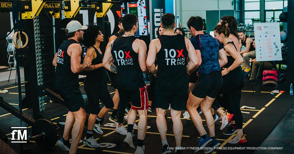 Mehrere Personen bilden einen Halbkreis im Fitnessstudio, sie tragen fast alle 10XTraining ärmellose Gym-Tops.