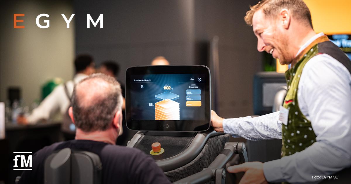 Ein Mann sitzt an einem EGYM-Gerät mit großem Bildschirm, ein weiterer Mann erklärt die Anzeige.