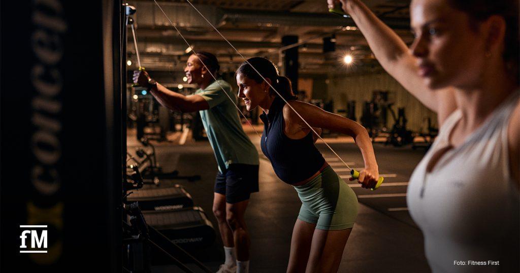 Drei Personen trainieren an Kabelzuggeräten in einem Fitnessstudio; im Vordergrund eine Frau, im Hintergrund zwei weitere Personen.