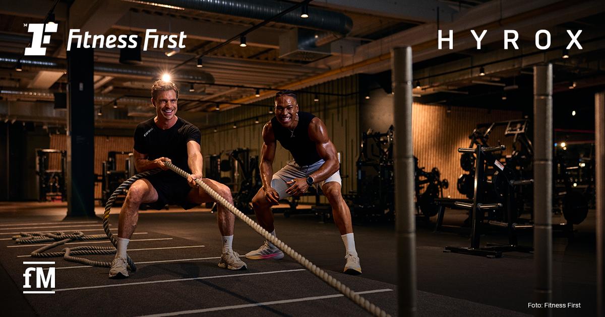 Zwei Männer trainieren mit Battle Ropes in einem Fitnessstudio; im Hintergrund Fitnessgeräte, Logos von Fitness First und HYROX eingeblendet.