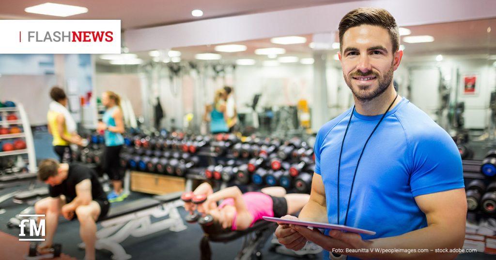 Mann im blauen Shirt mit Klemmbrett steht in Fitnessstudio, im Hintergrund trainieren mehrere Personen mit Hanteln.