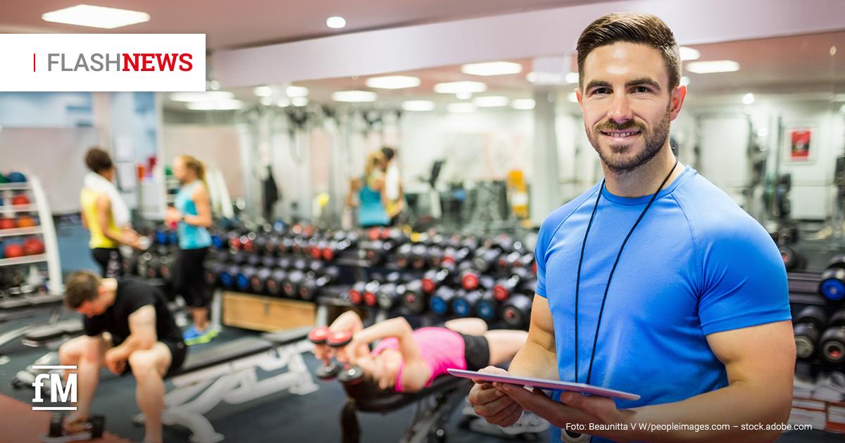 Mann im blauen Shirt mit Klemmbrett steht in Fitnessstudio, im Hintergrund trainieren mehrere Personen mit Hanteln.