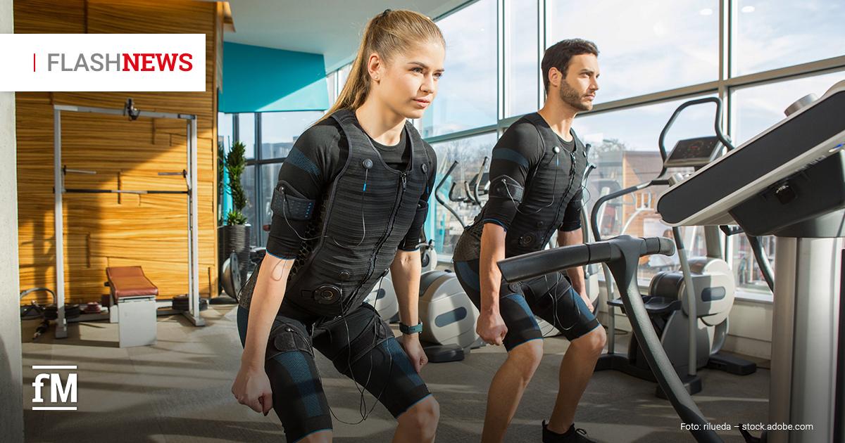 Zwei Personen, eine Frau und ein Mann, trainieren in einem Fitnessstudio mit Elektro-Muskelstimulationsanzügen; sie stehen in leichter Kniebeuge neben einem Laufband