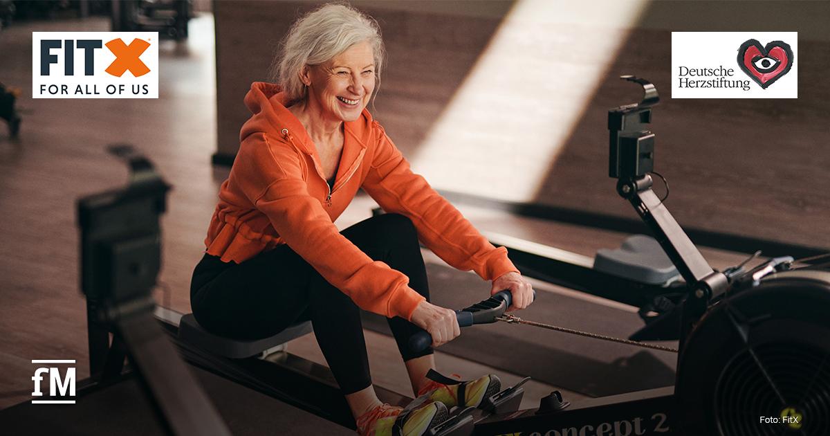 Eine ältere Frau mit grauen Haaren trainiert lachend an einem Rudergerät im Fitnessstudio. Im Hintergrund sind die Logos von FitX und der Deutschen Herzstiftung zu sehen.
