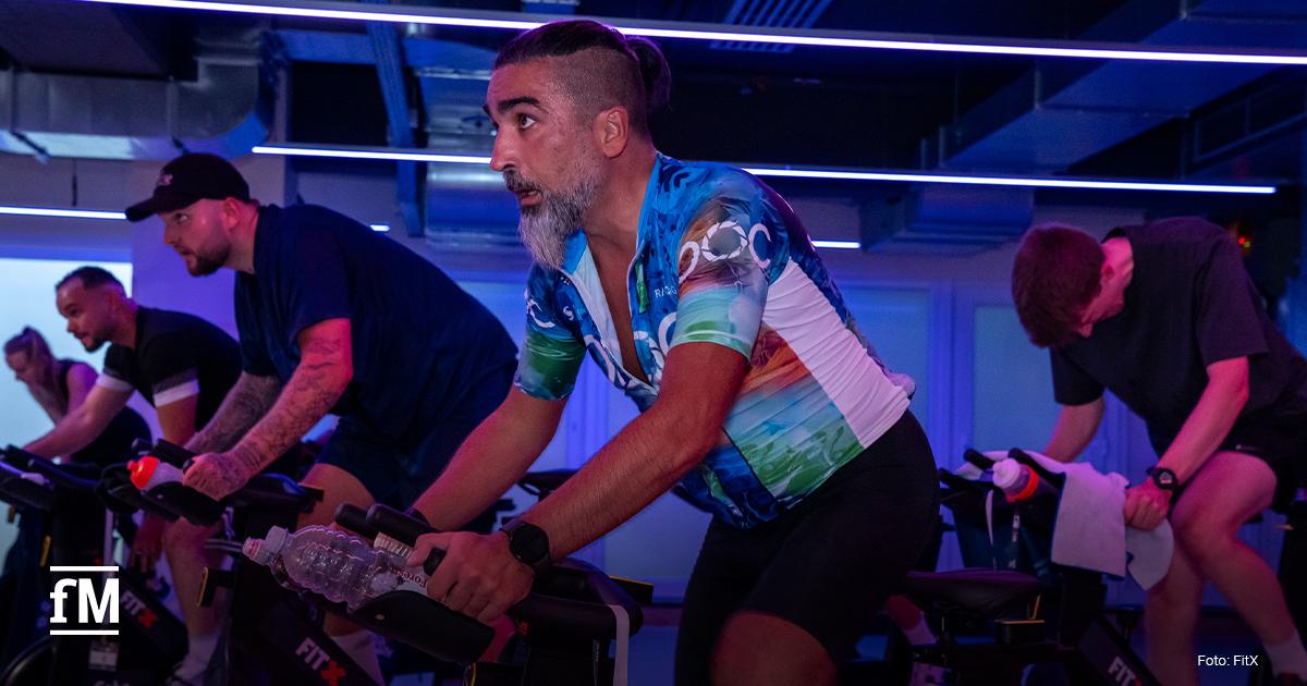 Mehrere Männer trainieren intensiv in einem abgedunkelten Raum auf Indoor-Cycling-Bikes. Im Vordergrund fährt ein Mann mit grauem Bart im bunten Trikot konzentriert.
