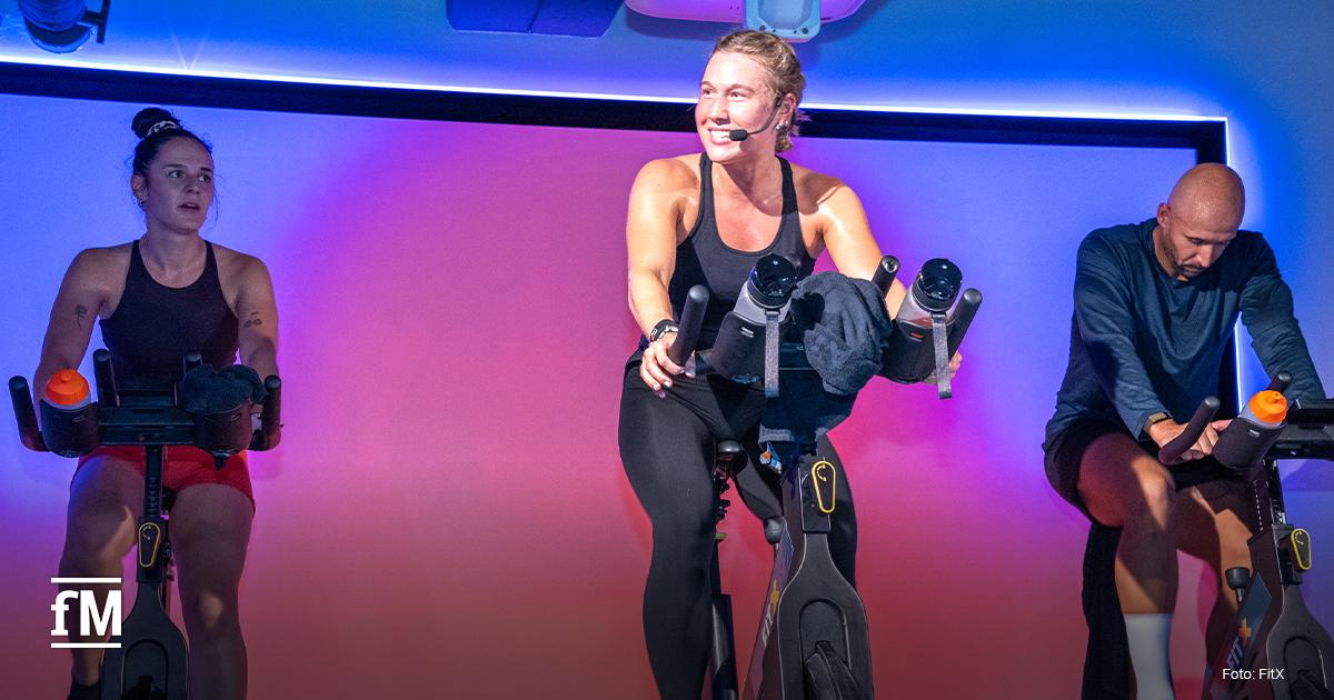 Eine Frau mit Headset motiviert als Trainerin zwei Teilnehmende während eines Indoor-Cycling-Kurses. Der Raum ist farbig beleuchtet, die Stimmung wirkt energiegeladen.