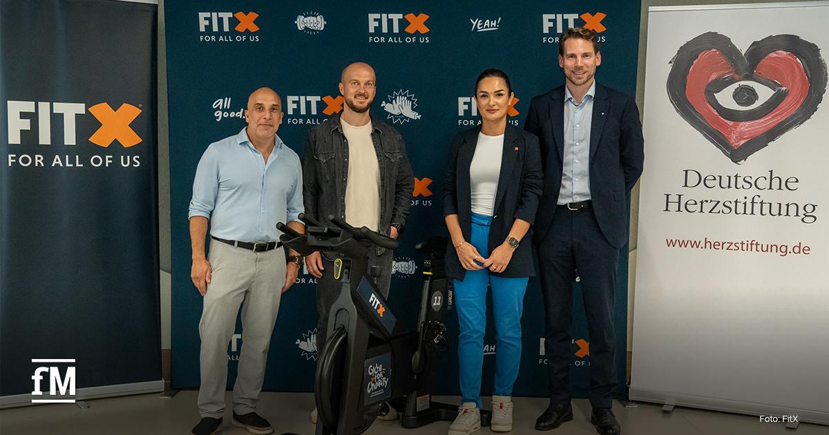 Vier Personen stehen neben einem Indoor-Bike vor Bannern von FitX und der Deutschen Herzstiftung. Sie präsentieren die Aktion 'Cycle for Charity'.