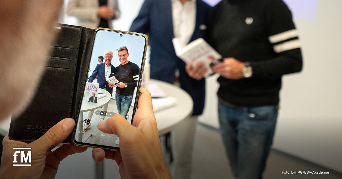 Eine Person fotografiert mit dem Smartphone zwei Männer, die gemeinsam posieren und jeweils ein Exemplar des Buches 'Lebenselixier Bewegung' in den Händen halten.