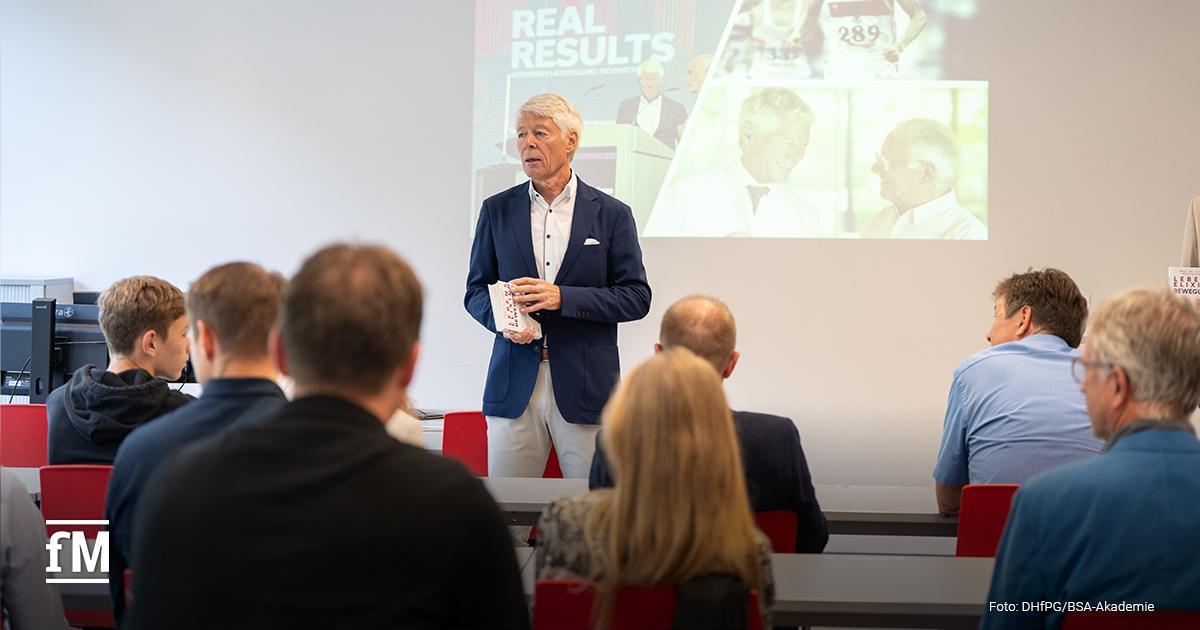 Prof. Dr. Thomas Wessinghage steht vor einer Projektionsfläche und spricht zu einer Gruppe von Zuhörern in einem Seminarraum. In der Hand hält er sein Buch 'Lebenselixier Bewegung'.