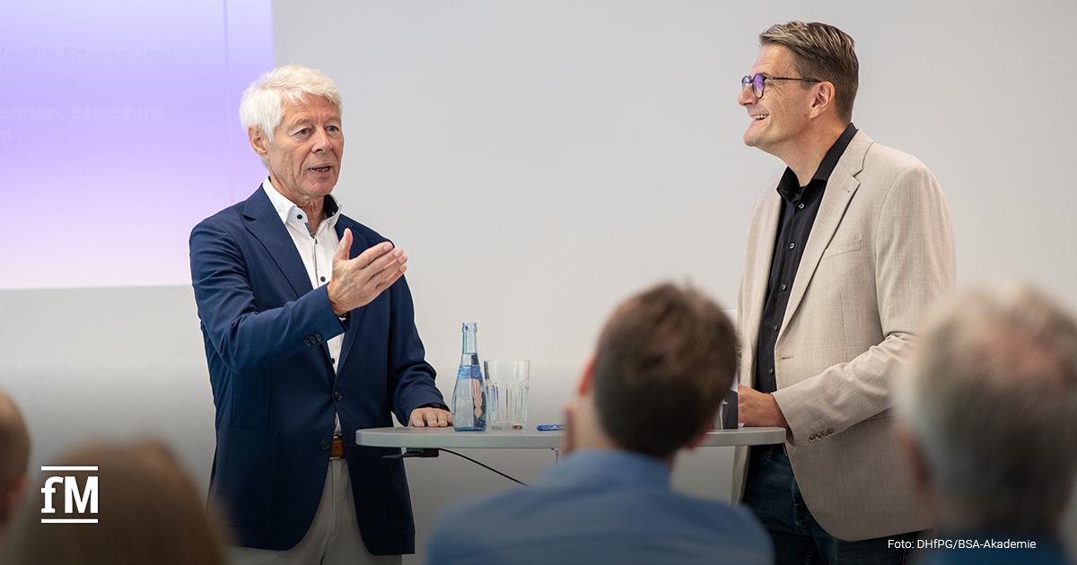 Prof. Dr. Thomas Wessinghage und ein Moderator stehen an einem Stehtisch und diskutieren vor Publikum. Wessinghage gestikuliert mit der Hand, während der Moderator lächelt.