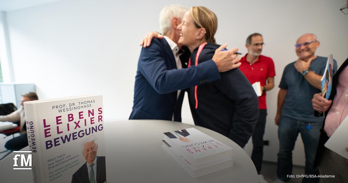 Prof. Dr. Thomas Wessinghage umarmt eine Frau (Sabine Kind) im Sport-Outfit während einer Signierstunde. Im Vordergrund liegt sein Buch auf dem Tisch.