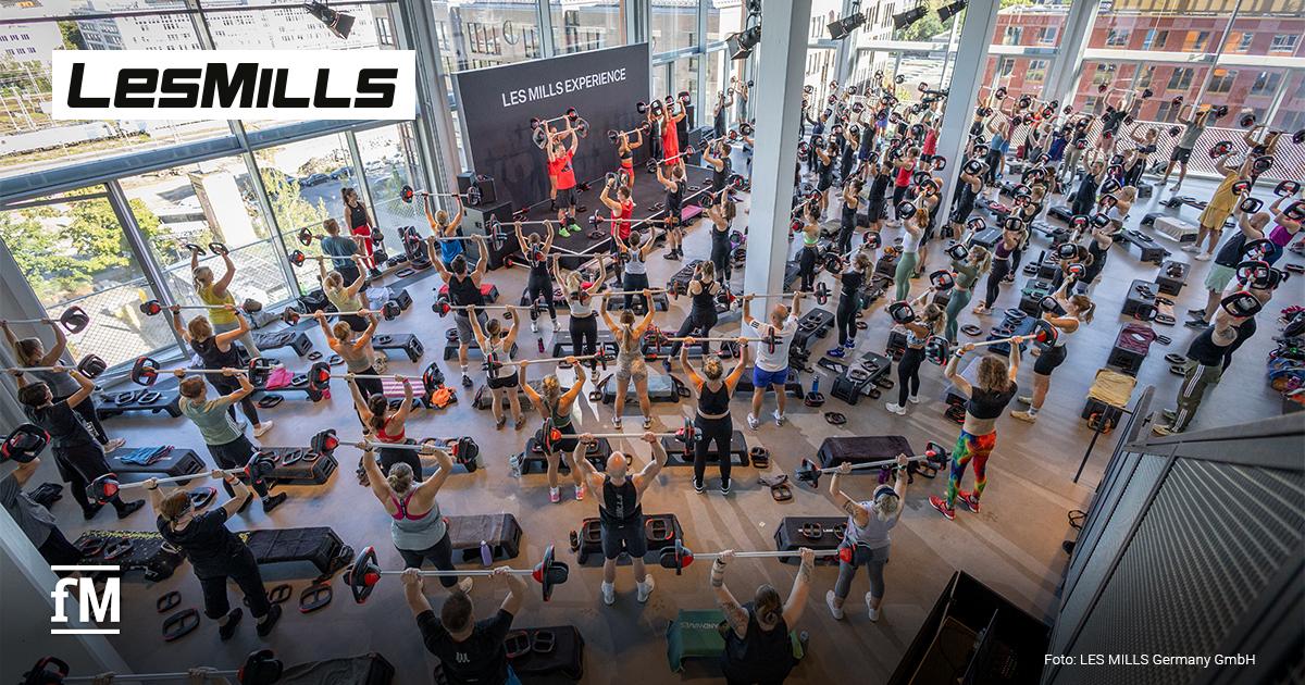 Große Gruppe trainiert mit Langhanteln vor Bühne mit Schriftzug „LES MILLS EXPERIENCE“