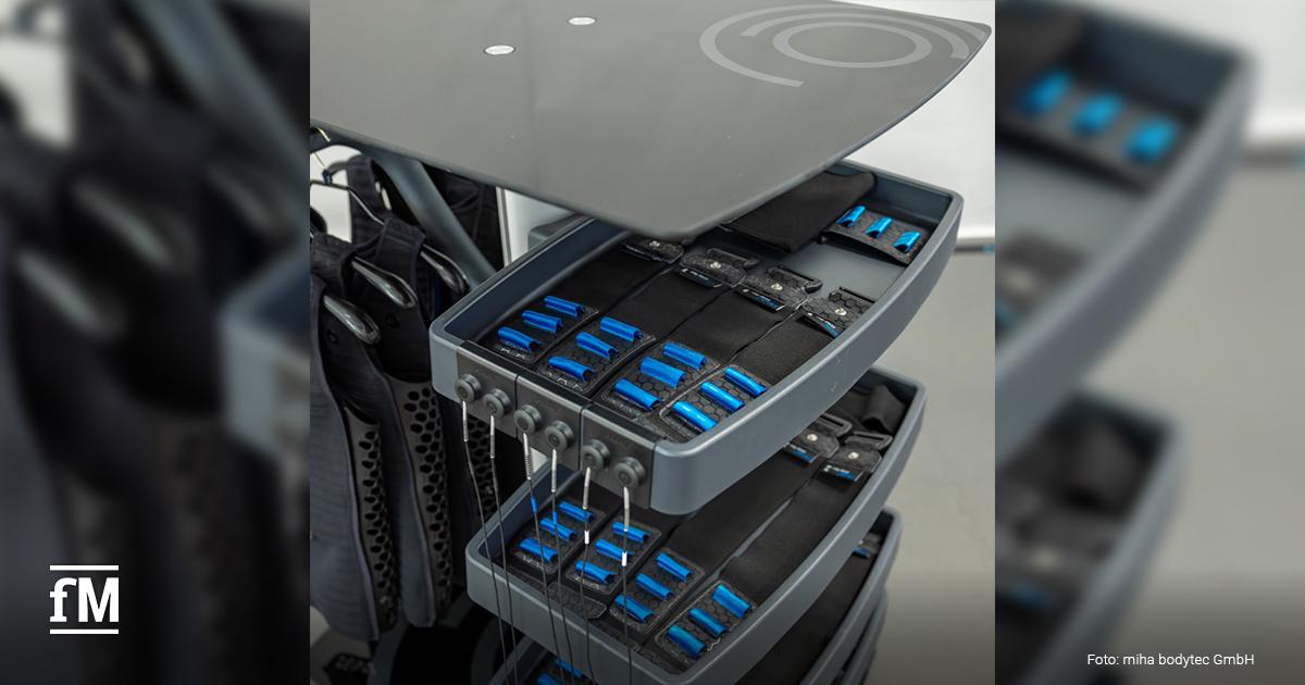 Mehrere Schubladen eines EMS-Geräts sind mit Elektrodenmodulen und Kabeln ausgestattet. Die Module sind ordentlich sortiert. Im Hintergrund hängen schwarze Anzüge für die Anwendung bereit