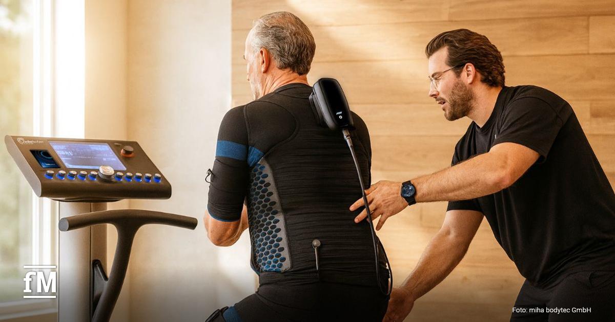 Ein Trainer unterstützt einen älteren Mann bei der Anwendung eines EMS-Trainingsgeräts. Der Mann trägt einen speziellen Anzug mit Elektroden, während der Trainer die Position am Rücken korrigiert.