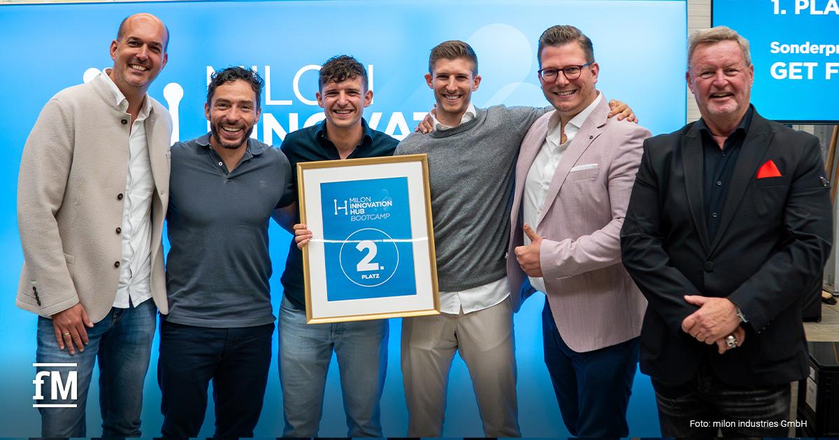 Gruppenfoto von sechs Personen auf der Bühne beim milon Innovation Hub Bootcamp. Zwei Männer in der Mitte halten eine Urkunde mit der Aufschrift '2. Platz SPORTABLE'. Alle lächeln vor blauem Hintergrund.