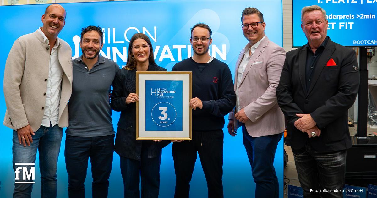 Gruppenfoto von sechs Personen beim milon Innovation Hub Bootcamp. Zwei Personen in der Mitte halten eine Urkunde mit der Aufschrift '3. Platz GET FIT'. Die Gruppe posiert vor blauem Hintergrund.