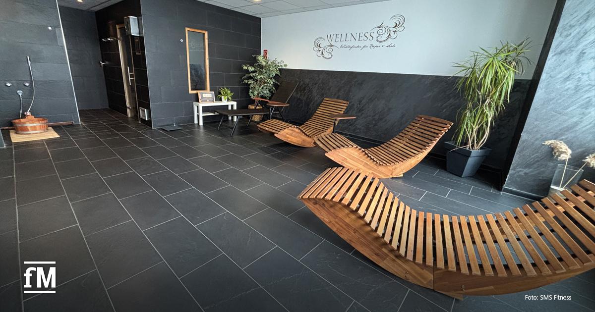 Heller Raum mit schwarzen Wänden, mehreren Holzliegen und grünen Pflanzen, an der Wand steht das Wort Wellness.