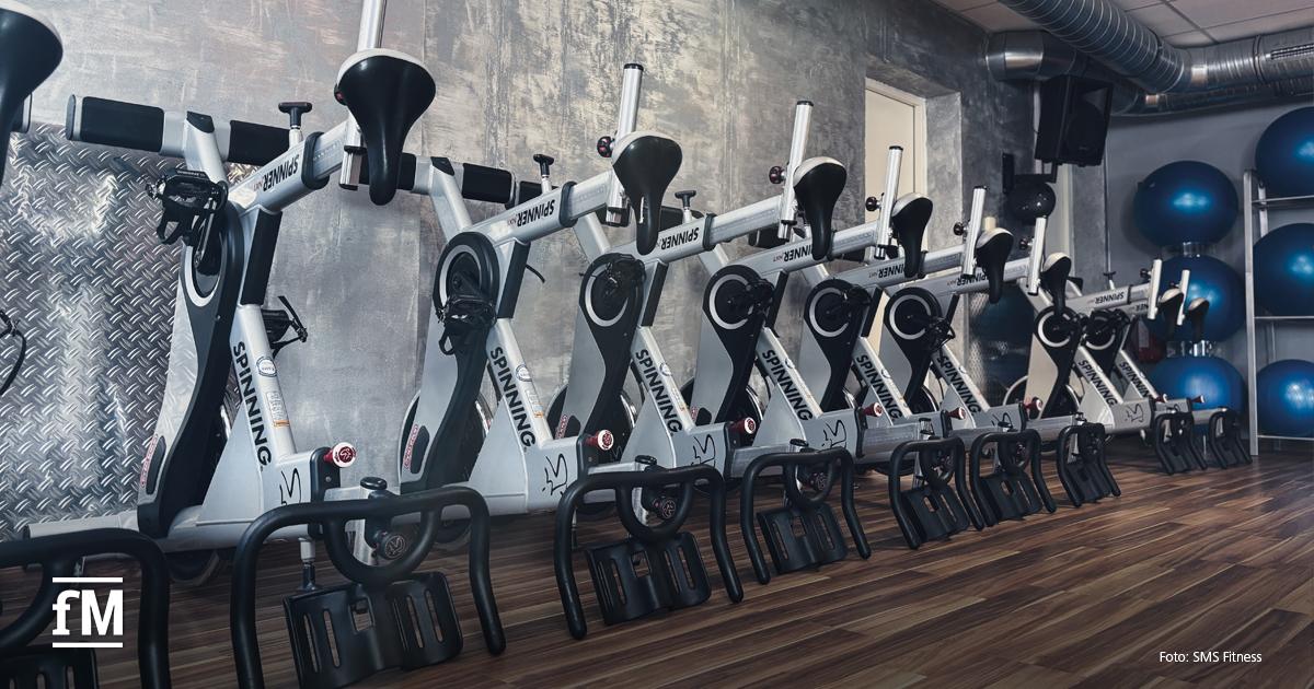 Mehrere Räder für Indoor Cycling in einer Reihe vor grauer Wand mit Holzboden und Gymnastikbällen im Hintergrund.
