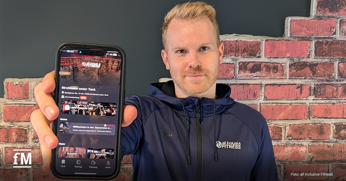 Ein Mann (Nico Miller) in dunkelblauer Fitnessjacke steht vor einer Wand mit Ziegelmuster und hält ein Smartphone nach vorn, auf dem eine Fitnessstudio-App mit dem Logo 'all inclusive Fitness' zu sehen ist.