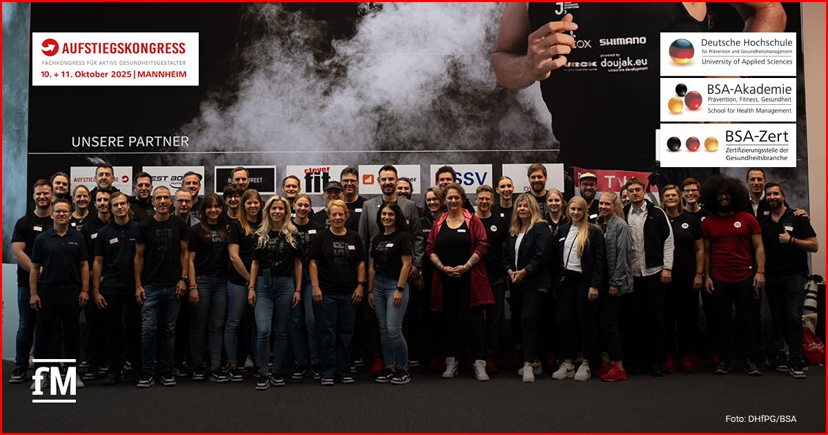 Eine große Gruppe von Menschen steht lächelnd vor einer Sponsorenwand mit Logos verschiedener Partner. Alle tragen dunkle Kleidung und Namensschilder, im Hintergrund ist ein Sportmotiv mit Rauchwolke zu sehen