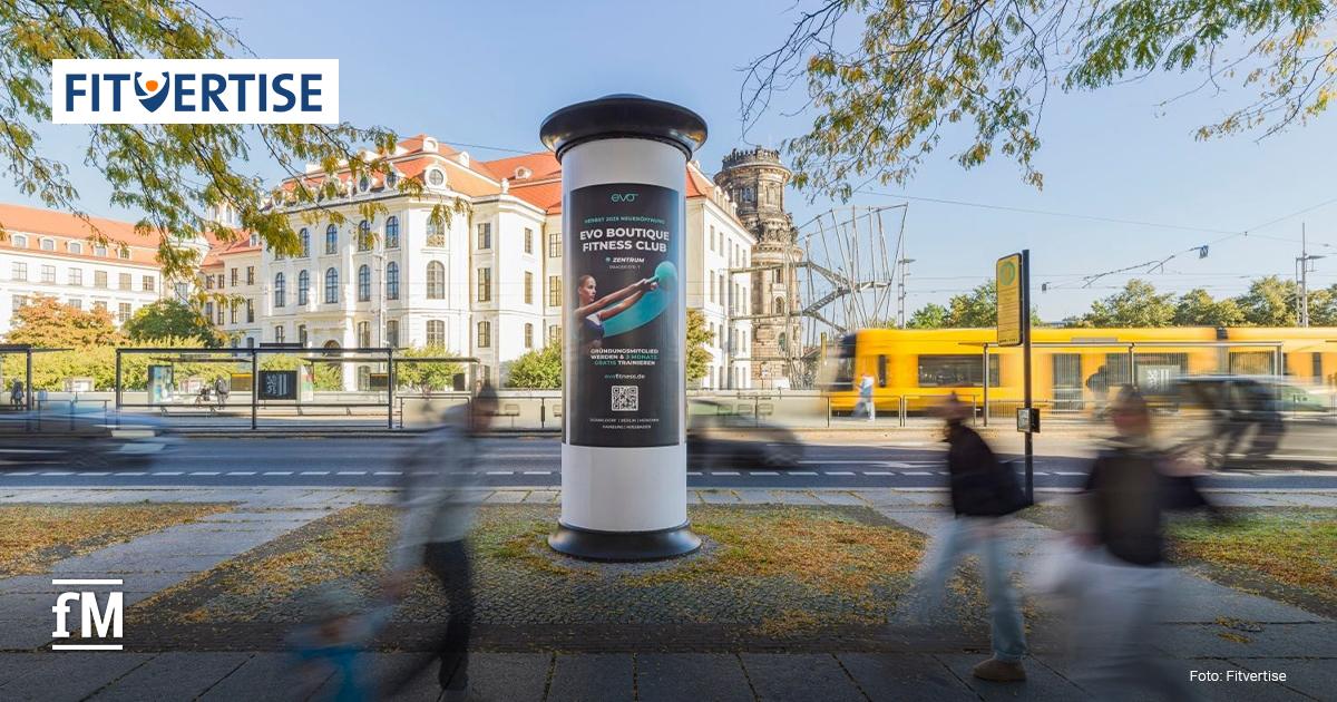 Litfaßsäule mit Werbung für ein Fitnessstudio, an einer Straße mit vorbeifahrender Straßenbahn und Passanten, im Stadtzentrum.