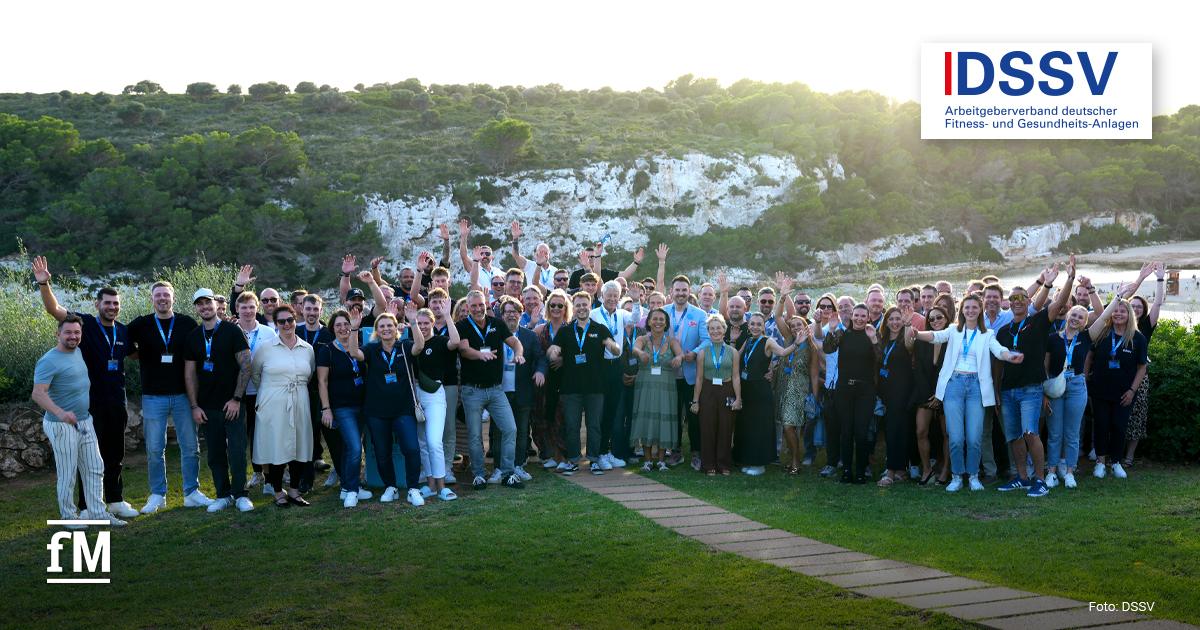 DSSV-Event Mallorca 2025 war ein voller Erfolg Eine große Gruppe von Menschen steht im Freien und posiert lachend für ein Gruppenfoto vor einer Küstenlandschaft mit Felsen und grünen Bäumen.