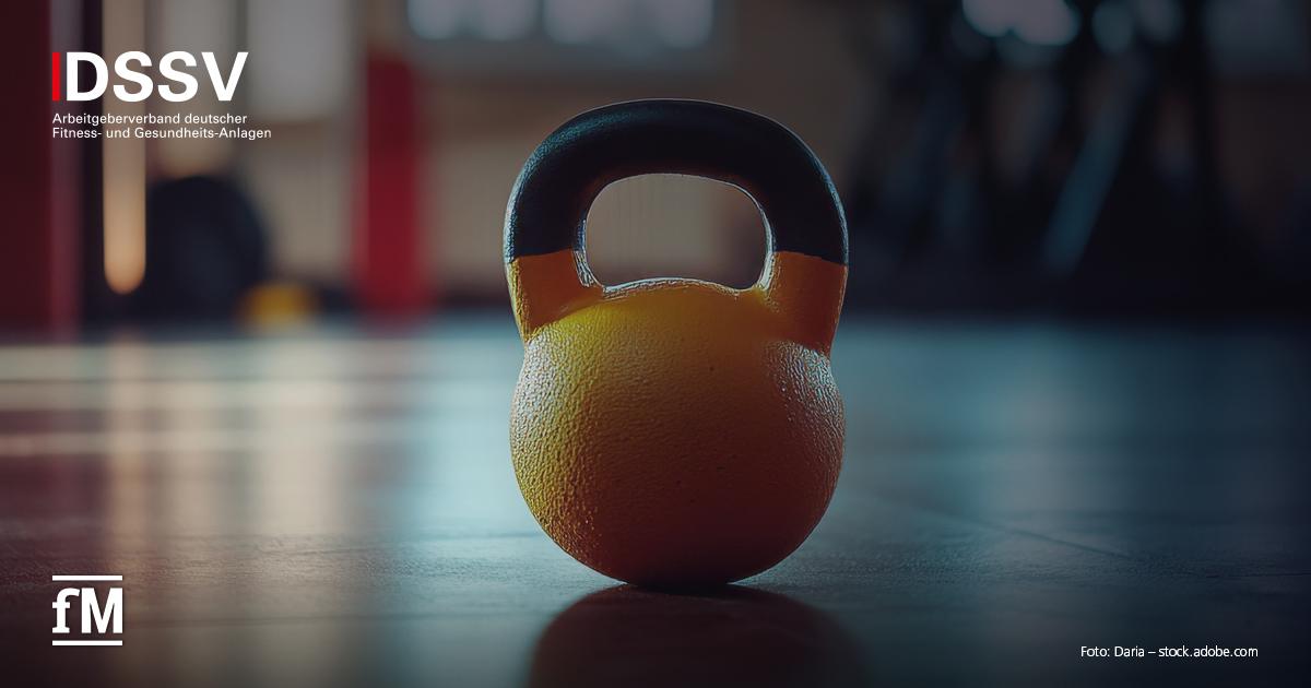 Gelbe Kettlebell mit schwarzem Griff auf Studioboden, im Hintergrund unscharfe Trainingsgeräte und Lichtreflexe.