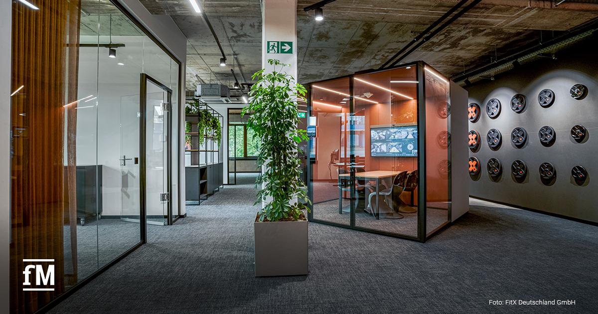 Büroflur mit Glaswänden, Pflanzen und einem orangefarbenen Besprechungsraum
