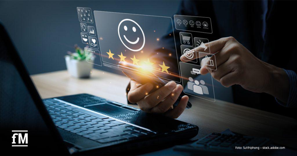Eine Person nutzt Smartphone und Laptop, während digitale Symbole wie ein Smiley mit fünf Sternen, Einkaufswagen und Chat-Icons holografisch über dem Gerät erscheinen – Symbol für Kundenzufriedenheit und digitales Feedback.