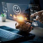 Eine Person nutzt Smartphone und Laptop, während digitale Symbole wie ein Smiley mit fünf Sternen, Einkaufswagen und Chat-Icons holografisch über dem Gerät erscheinen – Symbol für Kundenzufriedenheit und digitales Feedback.