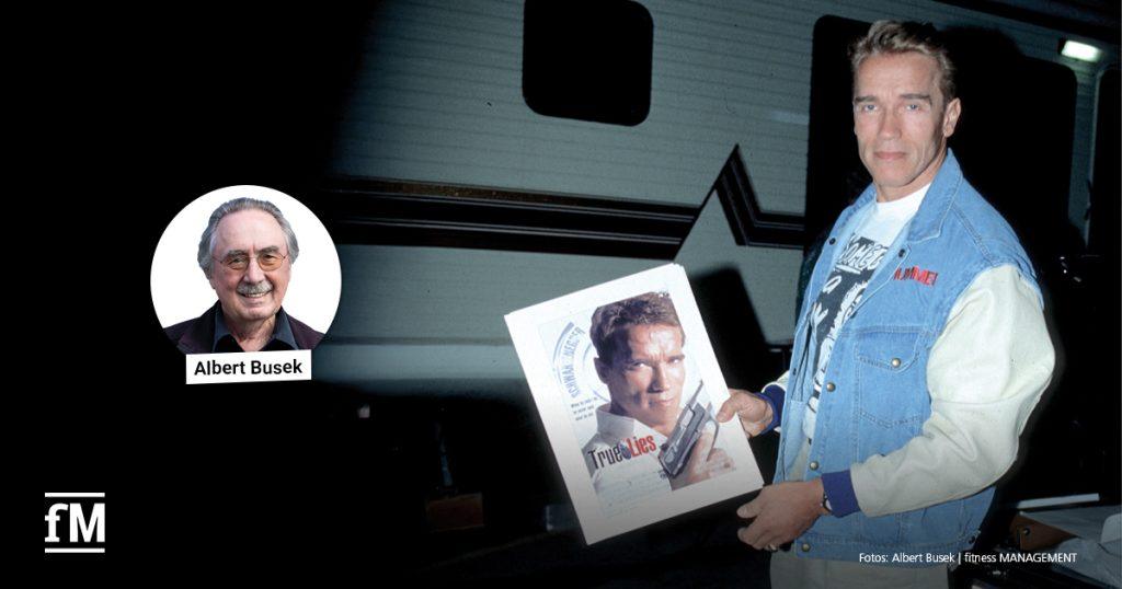 Arnold Schwarzenegger hält ein Filmplakat zu 'True Lies'; links im Bild ein Porträtfoto von Albert Busek mit Namensbeschriftung.