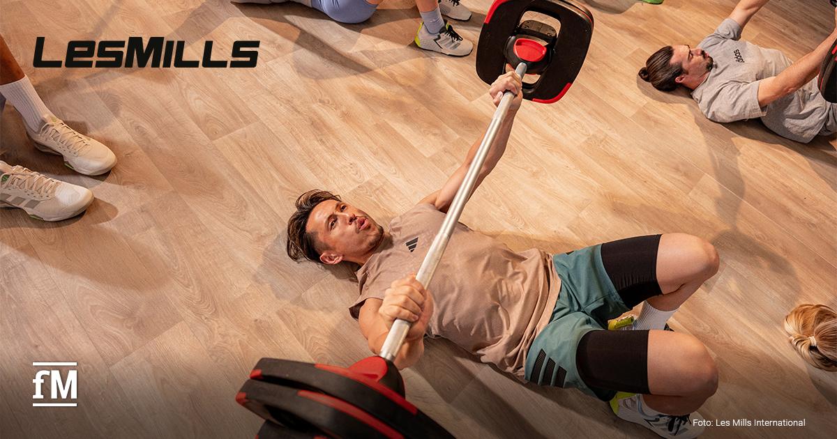 Ein Mann liegt auf einem hellen Parkettboden und drückt eine Langhantel nach oben, weitere Personen sind neben ihm zu erahnen, links oben das LES MILLS Logo in schwarz