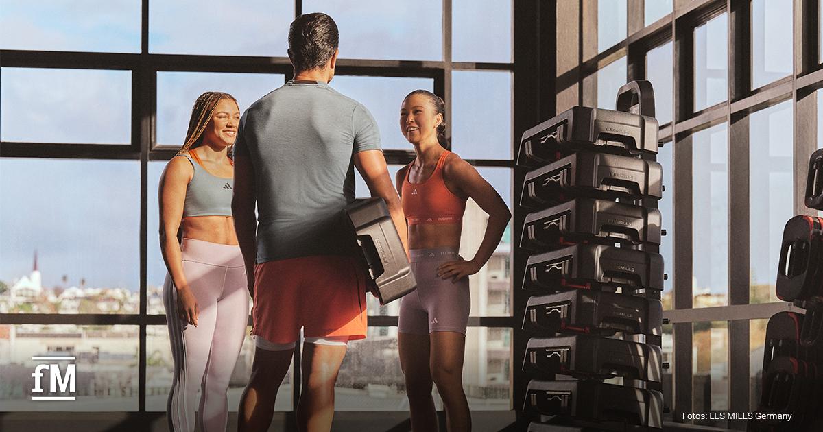 In einem hellen Fitnessstudio unterhalten sich drei Personen in Sportkleidung neben gestapelten Trainingsplattformen.