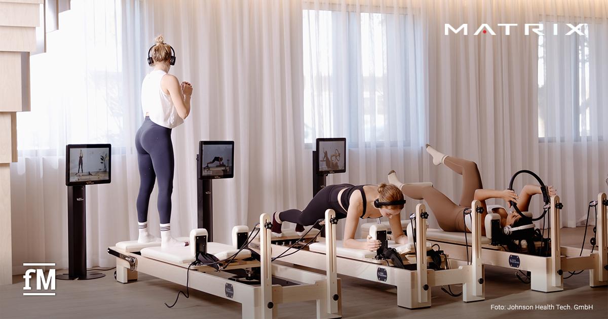 In einem hellen Studio trainieren drei Personen auf Reformer-Geräten mit Monitoren und Kopfhörern