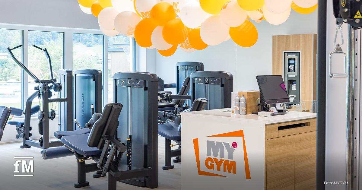 Empfangsbereich eines modernen Fitnessstudios mit Trainingsgeräten, Computerterminal und orange-weißen MYGYM-Luftballons an der Decke.