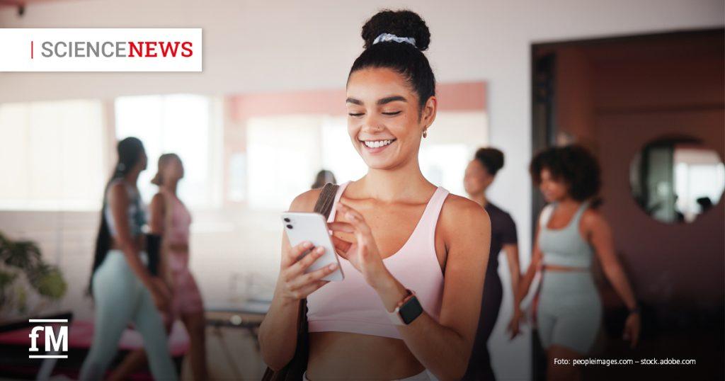 Junge Frau in Sportkleidung steht im Fitnessstudio und schaut lächelnd auf ihr Smartphone.