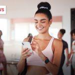 Junge Frau in Sportkleidung steht im Fitnessstudio und schaut lächelnd auf ihr Smartphone.