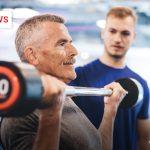 Älterer Mann hebt eine Hantel im Fitnessstudio, während ein Trainer ihn beobachtet.