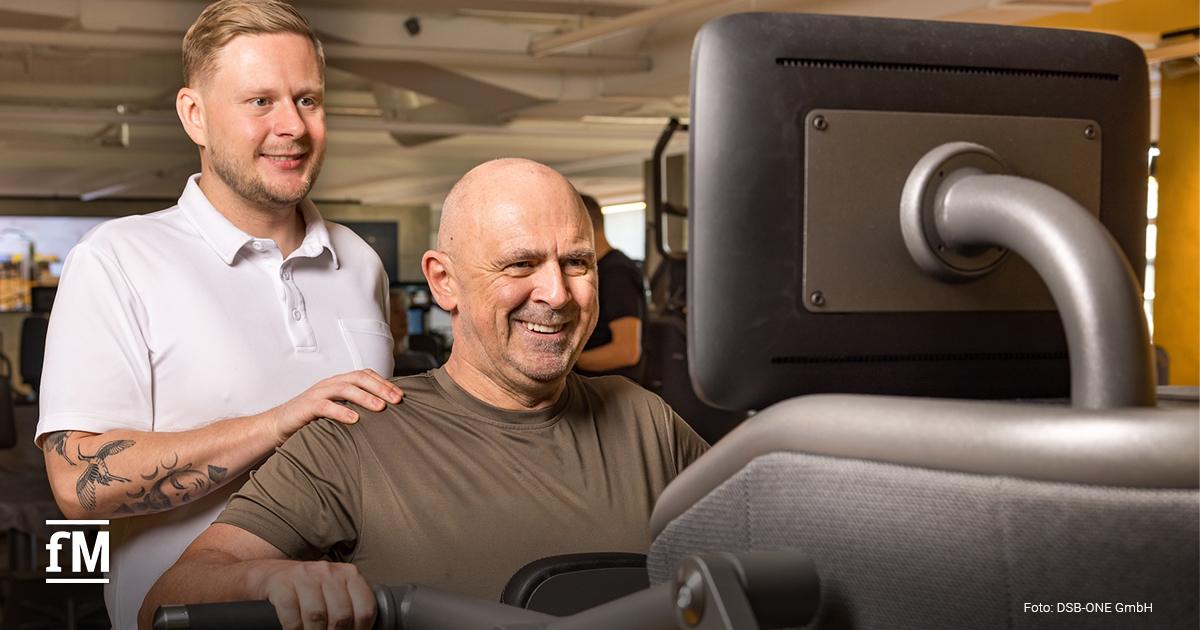 Ein Trainer in weißem Polo-Shirt unterstützt einen älteren Mann beim Training an einem Fitnessgerät mit Bildschirm. Beide wirken motiviert und freundlich.