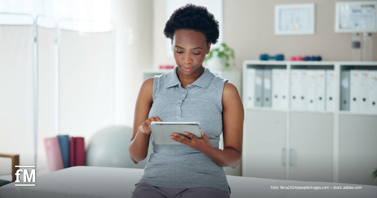 Frau sitzt auf Behandlungsliege und bedient Tablet in Praxisbüro mit Aktenschrank und Gymnastikball.