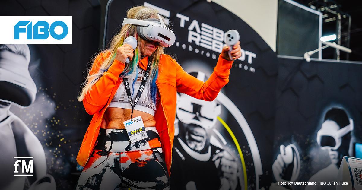 Eine blondhaarige Frau mittleren Alters trägt eine Virtual Reality-Brille und hält zwei Controller in den Händen, während sie in Boxhaltung vor einem Messestand auf der Fitnessmesse FIBO trainiert; im Hintergrund befinden sich großflächige Grafiken mit drei weiteren Personen in VR-Ausrüstung.