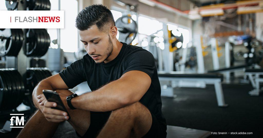 Ein Mann in Sportkleidung sitzt nach dem Training in einem Fitnessstudio auf dem Boden und schaut konzentriert auf sein Smartphone. Im Hintergrund sind Hanteln und Trainingsgeräte zu sehen.