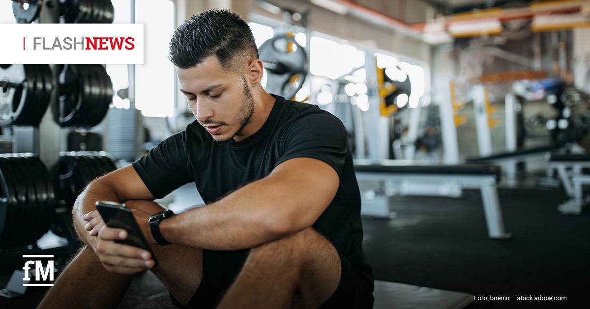 Ein Mann in Sportkleidung sitzt nach dem Training in einem Fitnessstudio auf dem Boden und schaut konzentriert auf sein Smartphone. Im Hintergrund sind Hanteln und Trainingsgeräte zu sehen.