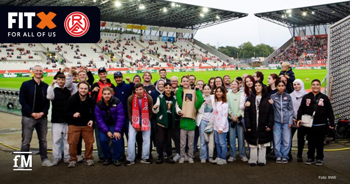 FitX, Rot-Weiss Essen und ESSENER CHANCEN Aktionsspieltag Eine große, bunt gemischte Gruppe junger Menschen posiert lächelnd am Spielfeldrand eines Stadions. Im Hintergrund sind die Tribünen mit einigen Zuschauern zu sehen. Oben links im Bild ist das Logo von FitX und Rot-Weiss Essen zu sehen.